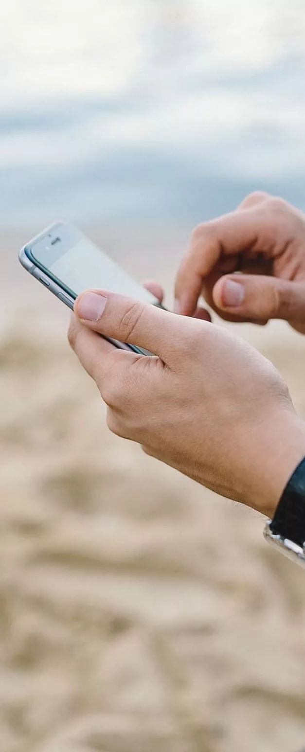 Ein Handy in den Händen eines Menschen der darauf tippt und im Hintergrund ein Strand.